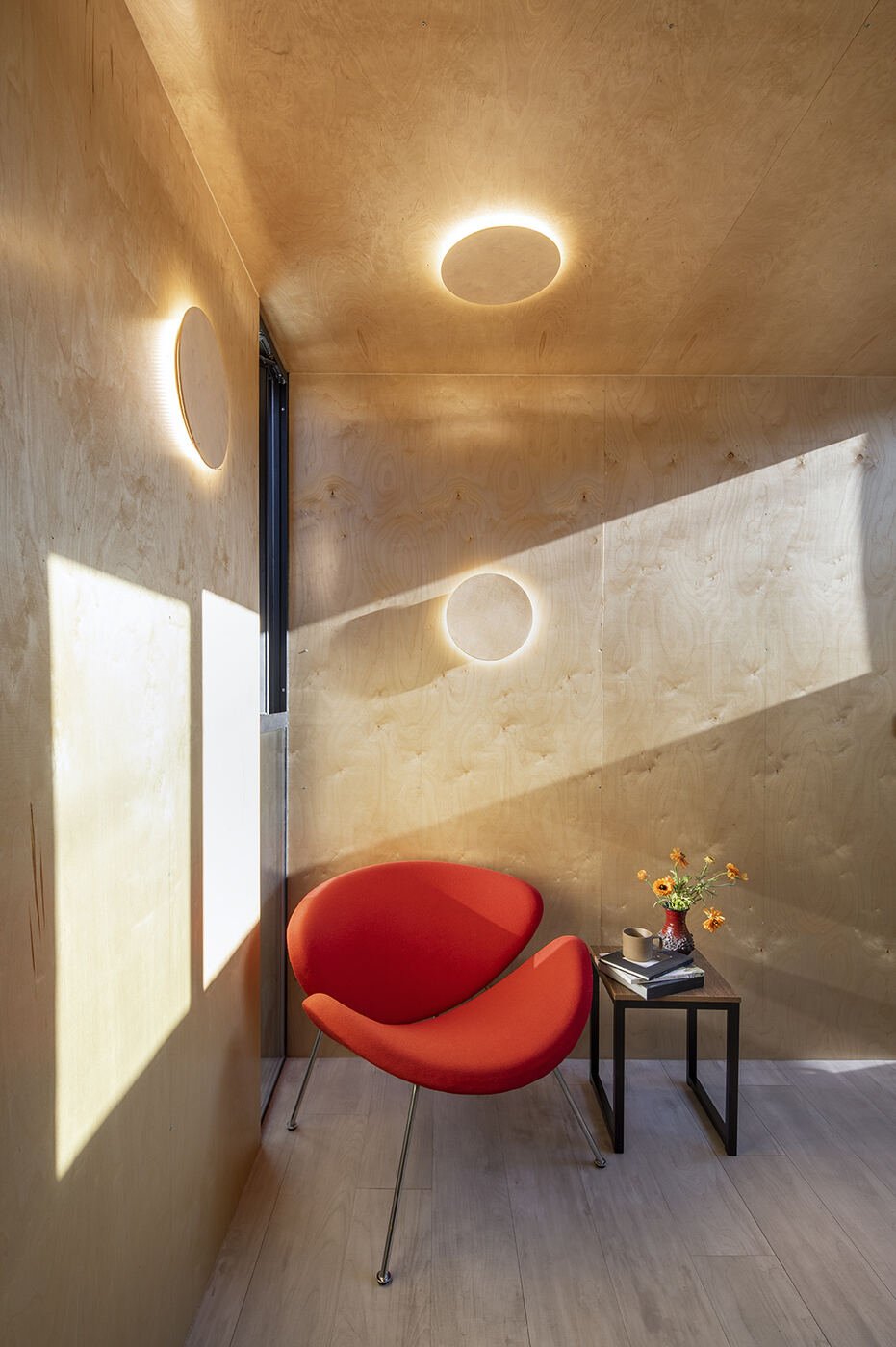 Plywood interior with round wall lights and a red lounge chair in afternoon sun
