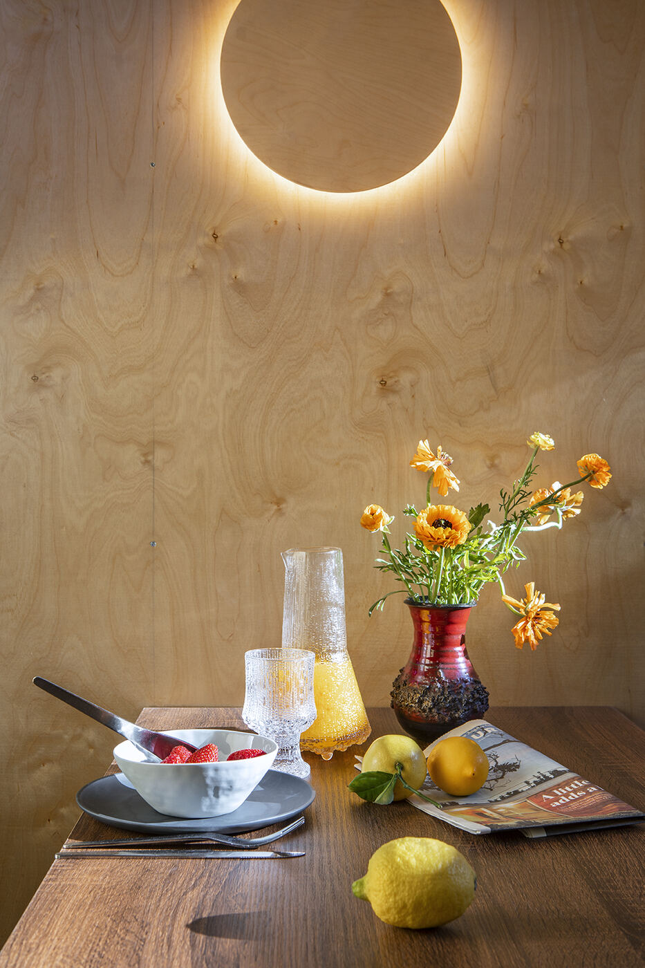 An eclipse-shaped light on a birch plywood wall above a dining table with flowers